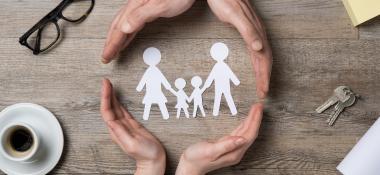 Top down view of a gray wood desktop that shows a pair of male hands and a pair of female hands surround a paper cutout of a family
